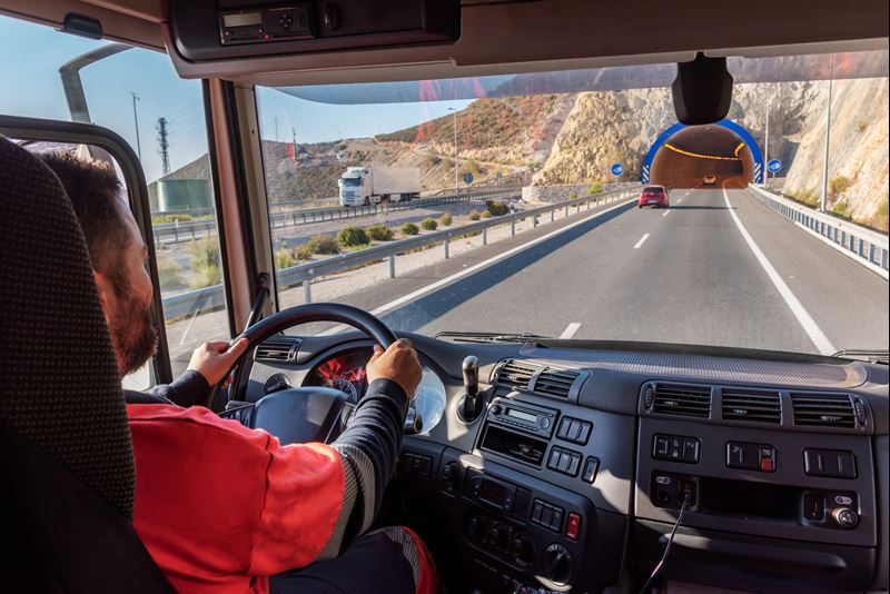 A truck driver, seen from inside the cab of his truck is sitting behind the steering wheel. He is driving on a highway. His truck is approaching the entrance of a tunnel.